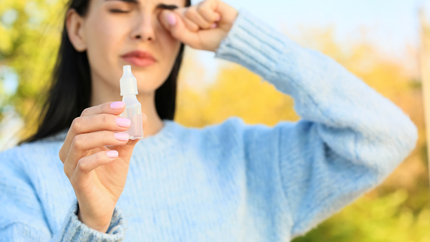 Woman rubbing her eyes due to contagious pink eye symptoms holding a bottle of eye drops