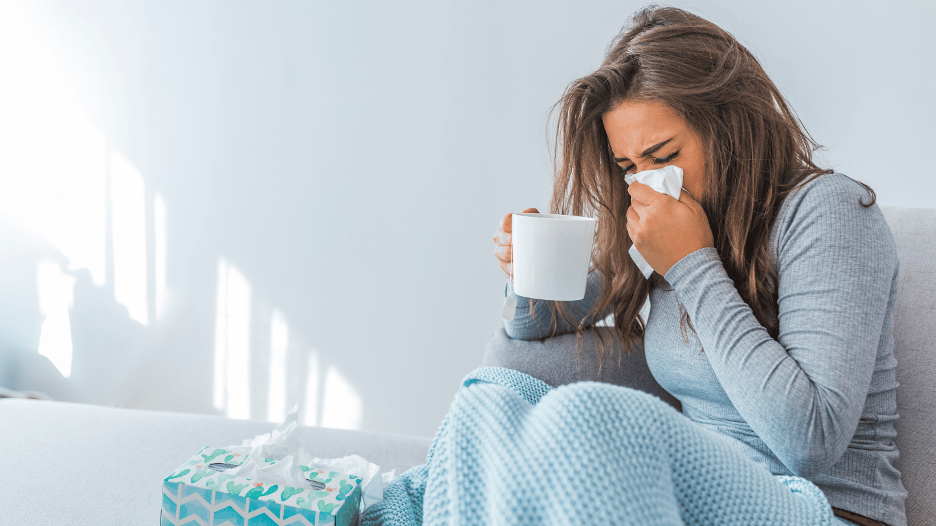 Woman with a cold blowing her nose while holding a mug, surrounded by tissues, highlighting the contagiousness of colds