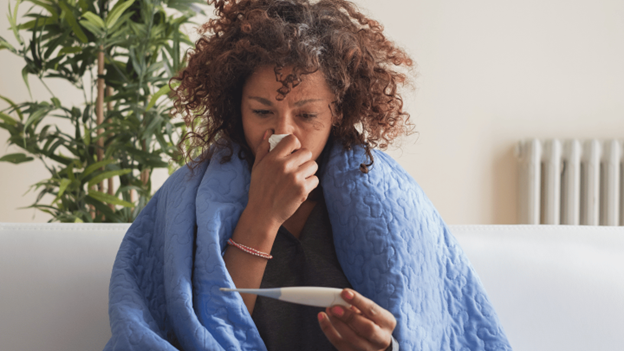 A woman wrapped in a blue blanket checks her thermometer while holding a tissue to her nose, showing signs of the severe flu from the 2025 flu season