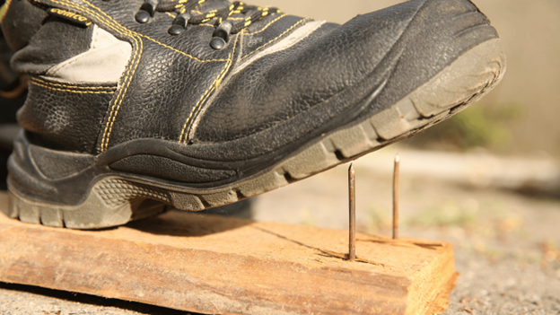 Work boot stepping on nails in a wooden board, illustrating the risk of puncture wounds and foot injuries