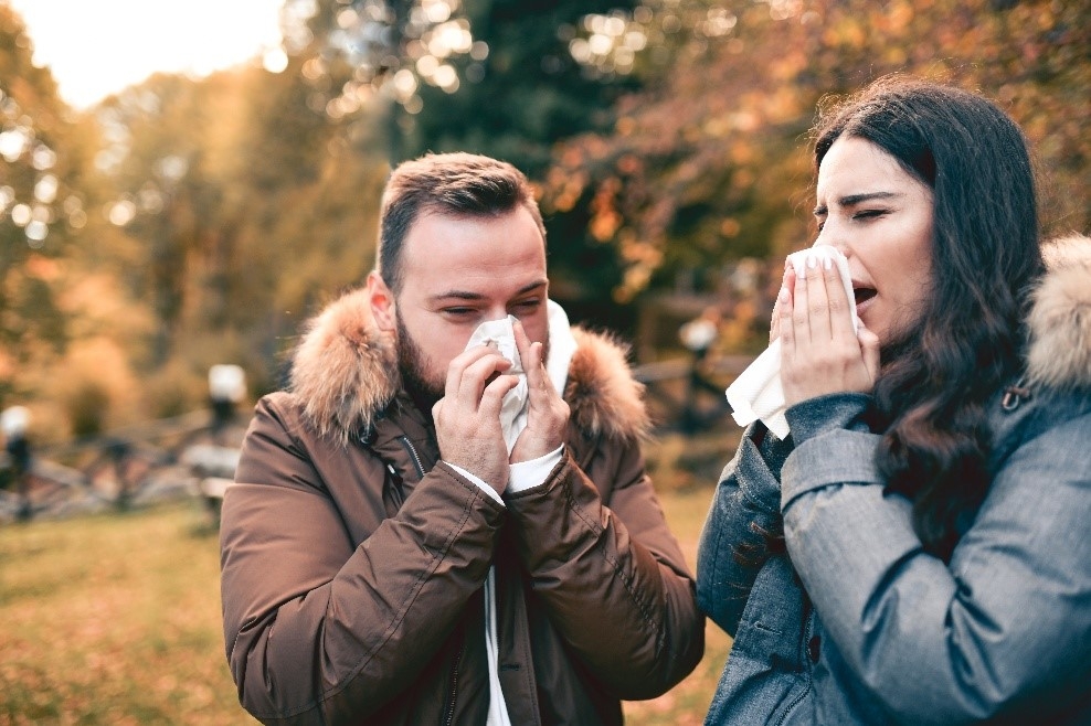 Two adults outdoors in winter holding tissues and showing flu symptoms.